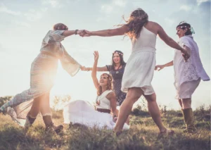 Eine Gruppe von Frauen genießt gemeinsam eine Auszeit in der Natur, umgeben von einem Feld bei Sonnenuntergang. Sie halten sich an den Händen, tanzen und lachen in einem Kreis, während eine von ihnen sich in der Mitte dreht. Die Szene strahlt Freude, Freiheit und Gemeinschaft aus, während jede Frau ihren eigenen Weg der Selbstfindung geht. Ihre Kleidung und Accessoires, von Blumenkränzen bis zu luftigen Kleidern, unterstreichen die ungezwungene und spirituelle Atmosphäre dieses besonderen Moments der Verbindung und des persönlichen Wachstums.