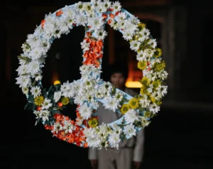 Ein Bild, das Kommunikation und Beziehung durch einen schwebenden Blumenkranz symbolisiert, der eine unsichtbare Verbindung zu einer schemenhaften Person im Hintergrund herstellt. Die Vielfalt der Blumen repräsentiert die Schönheit offener Kommunikation, die Beziehungen zum Erblühen bringt. Die Dunkelheit rundherum hebt diese Verbindung hervor und unterstreicht die Bedeutung gewaltfreier Kommunikation in zwischenmenschlichen Beziehungen