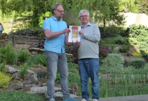 Zwei Herren stehen in einem sorgfältig gepflegten Garten, umgeben von einer Vielfalt an Pflanzen und einem ruhigen Teich. Sie halten gemeinsam ein Schild mit der Aufschrift 'TOP Medizin 2022', ein Symbol für Anerkennung und Erfolg. Diese Szene könnte metaphorisch für die Errungenschaften in der Psychotherapie stehen, bei der Zusammenarbeit, Engagement und die Pflege des geistigen Wohlbefindens zu anerkennenswerten Fortschritten führen. Der Garten symbolisiert das Wachstum und die Entwicklung, die in der therapeutischen Arbeit erreicht werden können, während die harmonische Umgebung die Wichtigkeit einer unterstützenden und beruhigenden Atmosphäre für den Heilungsprozess unterstreicht.