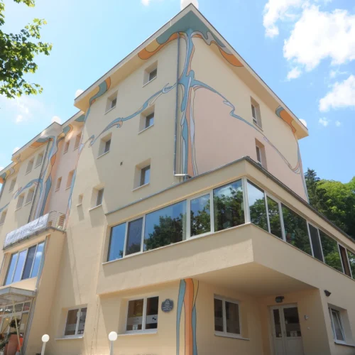 Das Gebäude der Akademie Heiligenfeld in Bad Kissingen, ein Seminarhaus mit auffälliger, kunstvoller Fassadengestaltung, steht sonnenbeschienen vor einem Hintergrund aus dichtem Grün und blauem Himmel.