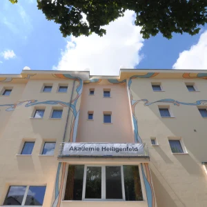 GPT Ein helles Gebäude der Akademie Heiligenfeld, spezialisiert auf Fort- und Weiterbildung, mit kunstvollen Verzierungen und einem klaren Himmel im Hintergrund.