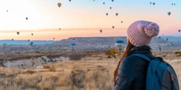 Eine Person steht in stiller Betrachtung vor einer atemberaubenden Kulisse, die von zahlreichen Heißluftballons in den frühen Morgenstunden belebt wird. Die sanften Farben des Sonnenaufgangs spiegeln sich in den Ballons wider, die friedlich über die einzigartige Landschaft schweben. Diese Szene symbolisiert einen Moment der Meditation und Selbstfindung, in dem die Person, umgeben von der majestätischen Schönheit der Natur, einen tiefen inneren Frieden und eine Verbindung zum eigenen Selbst erlebt. Die Weite des Himmels und die sanfte Bewegung der Ballons bieten eine perfekte Metapher für die Reise der Selbstentdeckung und die unendlichen Möglichkeiten, die sich öffnen, wenn man sich der Stille und der Reflexion hingibt