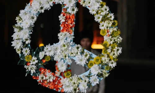 Ein Bild, das Kommunikation und Beziehung durch einen schwebenden Blumenkranz symbolisiert, der eine unsichtbare Verbindung zu einer schemenhaften Person im Hintergrund herstellt. Die Vielfalt der Blumen repräsentiert die Schönheit offener Kommunikation, die Beziehungen zum Erblühen bringt. Die Dunkelheit rundherum hebt diese Verbindung hervor und unterstreicht die Bedeutung gewaltfreier Kommunikation in zwischenmenschlichen Beziehungen