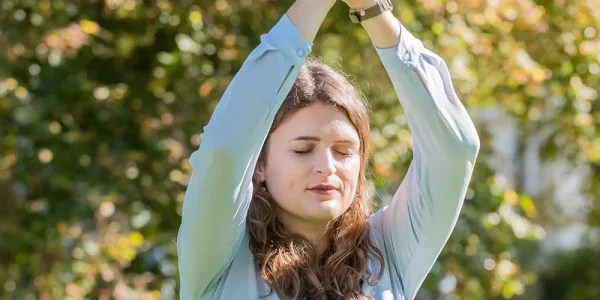 Eine junge Frau steht friedlich im sanften Licht eines herbstlichen Gartens, die Augen geschlossen, die Arme sanft über ihrem Kopf erhoben, als ob sie die Stille und die frische Luft um sich herum aufnimmt. Dieses Bild fängt einen Moment der Selbstreflexion und Ruhe ein, symbolisch für die tiefe innere Welt und die Empfänglichkeit hochsensibler Personen gegenüber ihrer Umgebung. Die natürliche Kulisse und das weiche Sonnenlicht unterstreichen die Harmonie und das Bedürfnis nach einem umsorgten Raum, in dem hochsensible Menschen ihre Sensibilität als Stärke erleben können.