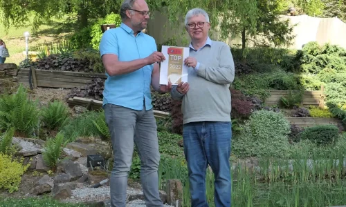 Zwei Herren stehen in einem sorgfältig gepflegten Garten, umgeben von einer Vielfalt an Pflanzen und einem ruhigen Teich. Sie halten gemeinsam ein Schild mit der Aufschrift 'TOP Medizin 2022', ein Symbol für Anerkennung und Erfolg. Diese Szene könnte metaphorisch für die Errungenschaften in der Psychotherapie stehen, bei der Zusammenarbeit, Engagement und die Pflege des geistigen Wohlbefindens zu anerkennenswerten Fortschritten führen. Der Garten symbolisiert das Wachstum und die Entwicklung, die in der therapeutischen Arbeit erreicht werden können, während die harmonische Umgebung die Wichtigkeit einer unterstützenden und beruhigenden Atmosphäre für den Heilungsprozess unterstreicht.