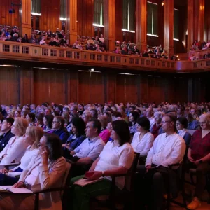 Ein aufmerksames Publikum sitzt in einem prachtvollen Saal, die Blicke gebannt auf die Bühne gerichtet, wo ein Vortrag im Rahmen eines Kongresses stattfindet. Die Teilnehmer verkörpern Offenheit und Bereitschaft zum Dialog und Wissensaustausch, eingetaucht in die inspirierende Atmosphäre des Kongresses.