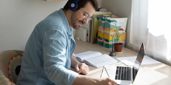 Ein engagierter Mann nutzt Online-Angebote und nimmt an einem Online-Seminar teil, während er von zu Hause aus arbeitet. Er trägt Kopfhörer und eine Brille, konzentriert sich auf Notizen, die er neben seinem Laptop macht, auf dem offensichtlich eine Lernplattform oder ein Webinar geöffnet ist. Um ihn herum sind Bücher und Unterlagen ausgebreitet, die seine Beteiligung und sein Engagement im Online-Lernen unterstreichen. Das natürliche Licht, das den Raum durchflutet, und die ruhige, organisierte Umgebung tragen zur Effektivität seines Studiums bei. Diese Szene verkörpert die moderne Bildung und die Anpassungsfähigkeit an digitale Lernumgebungen durch die Nutzung von Online-Angeboten und Seminaren.