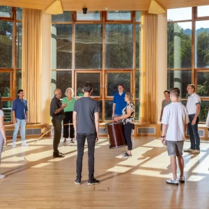 Eine Gruppe von Menschen steht in einem hellen, geräumigen Seminarraum mit großen Fenstern, die einen Blick auf die umgebende Natur freigeben. Sie sind in einer interaktiven Übung engagiert, geführt von einem Seminarleiter, was die dynamische Atmosphäre eines Bildungsseminars unterstreicht.