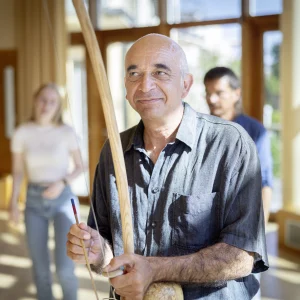 Ein Mann mit einem freundlichen Lächeln hält in einem hellen, offenen Raum mit großen Fenstern einen Stock und einen Schlegel, bereit, einen TaKeTiNa®-Rhythmus-Workshop zu leiten, während im Hintergrund Teilnehmende in Erwartung der nächsten Übung stehen.